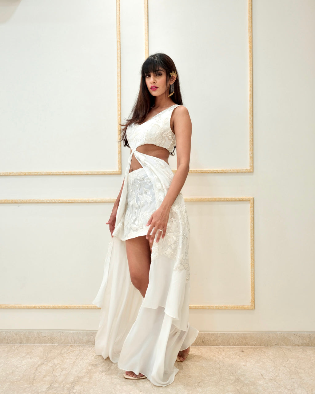 Woman wearing a white dress with a high slit in front of a white wall.