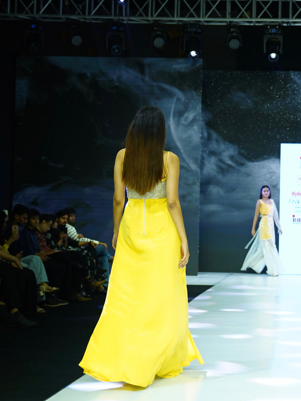 Model in a yellow dress walking on a fashion show runway with a dark backdrop.