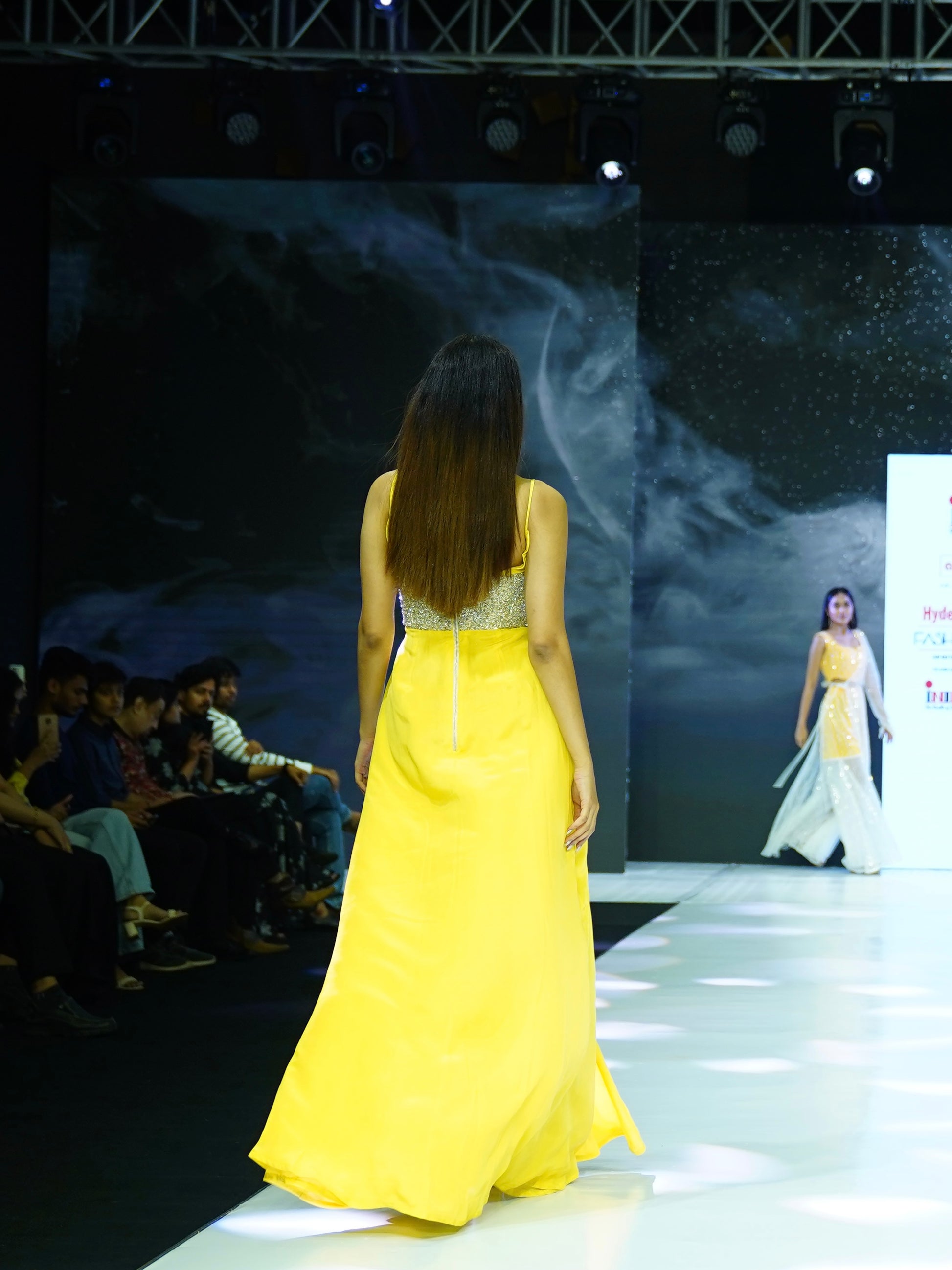 Model in a yellow dress walking on a fashion show runway with a dark backdrop.