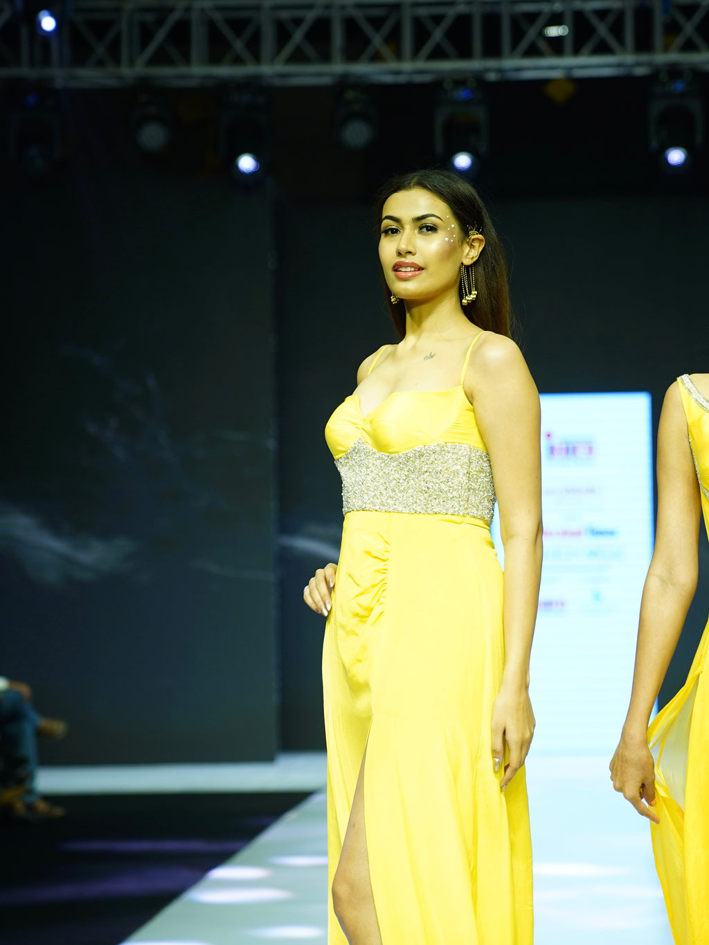 Model wearing a yellow evening gown with a slit on a runway.