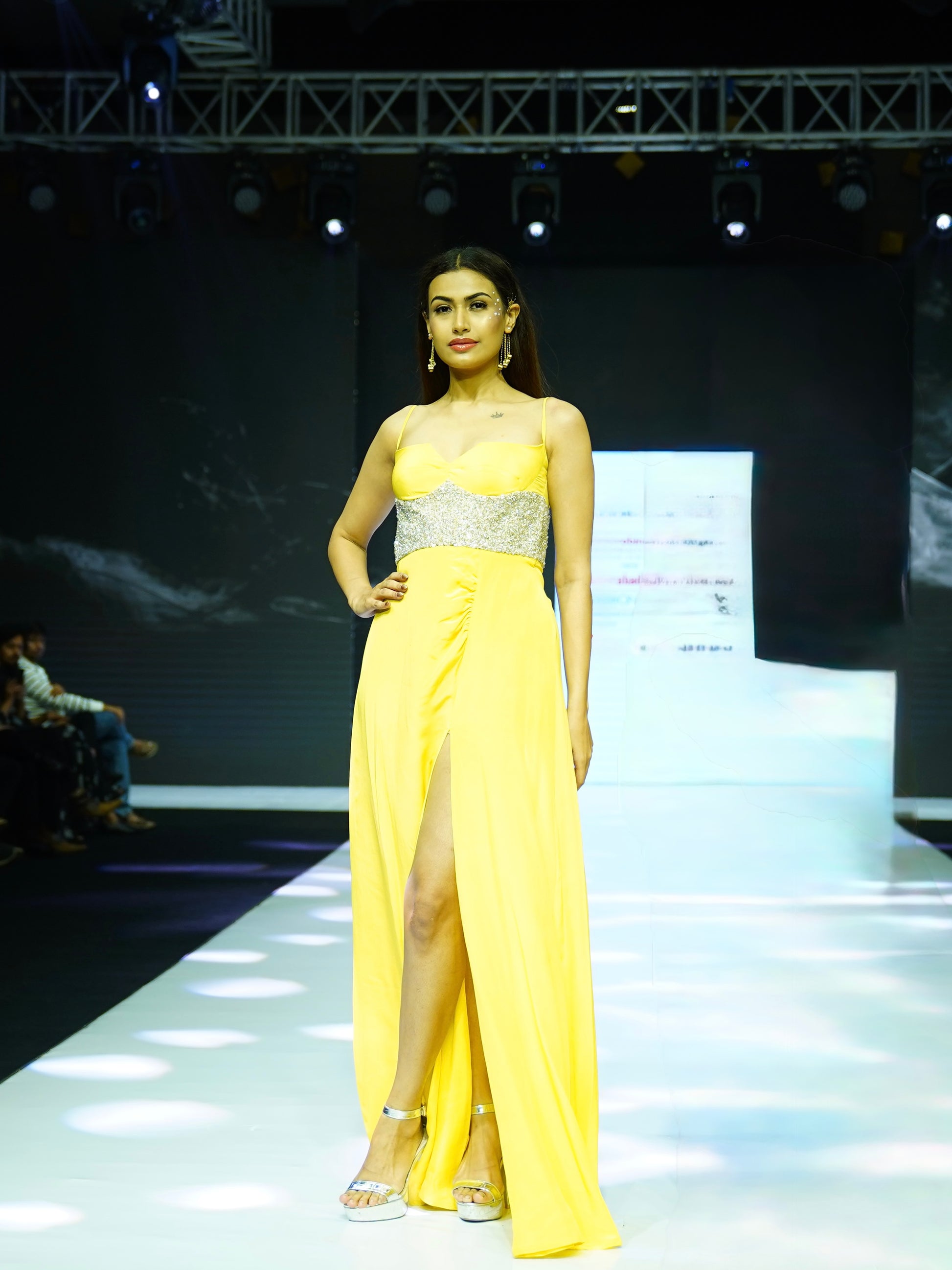 Model wearing a yellow evening gown with a silver belt on a runway.