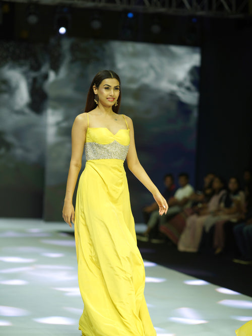 Model wearing a yellow dress on a fashion show runway with an audience in the background.