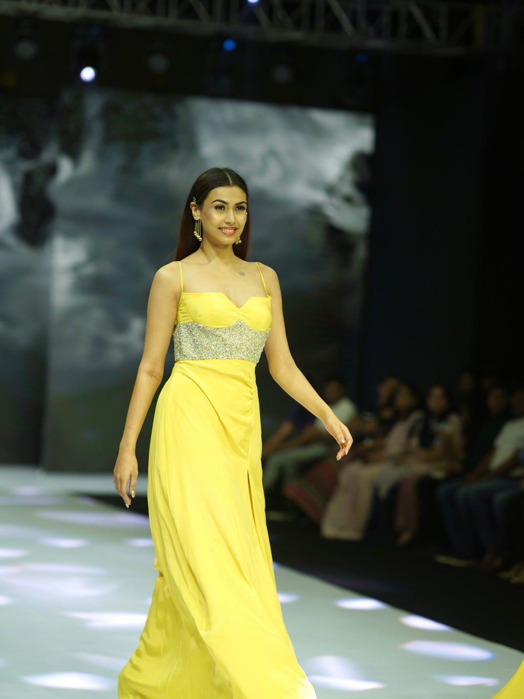 Model in a yellow evening gown with a silver top on a fashion show runway.