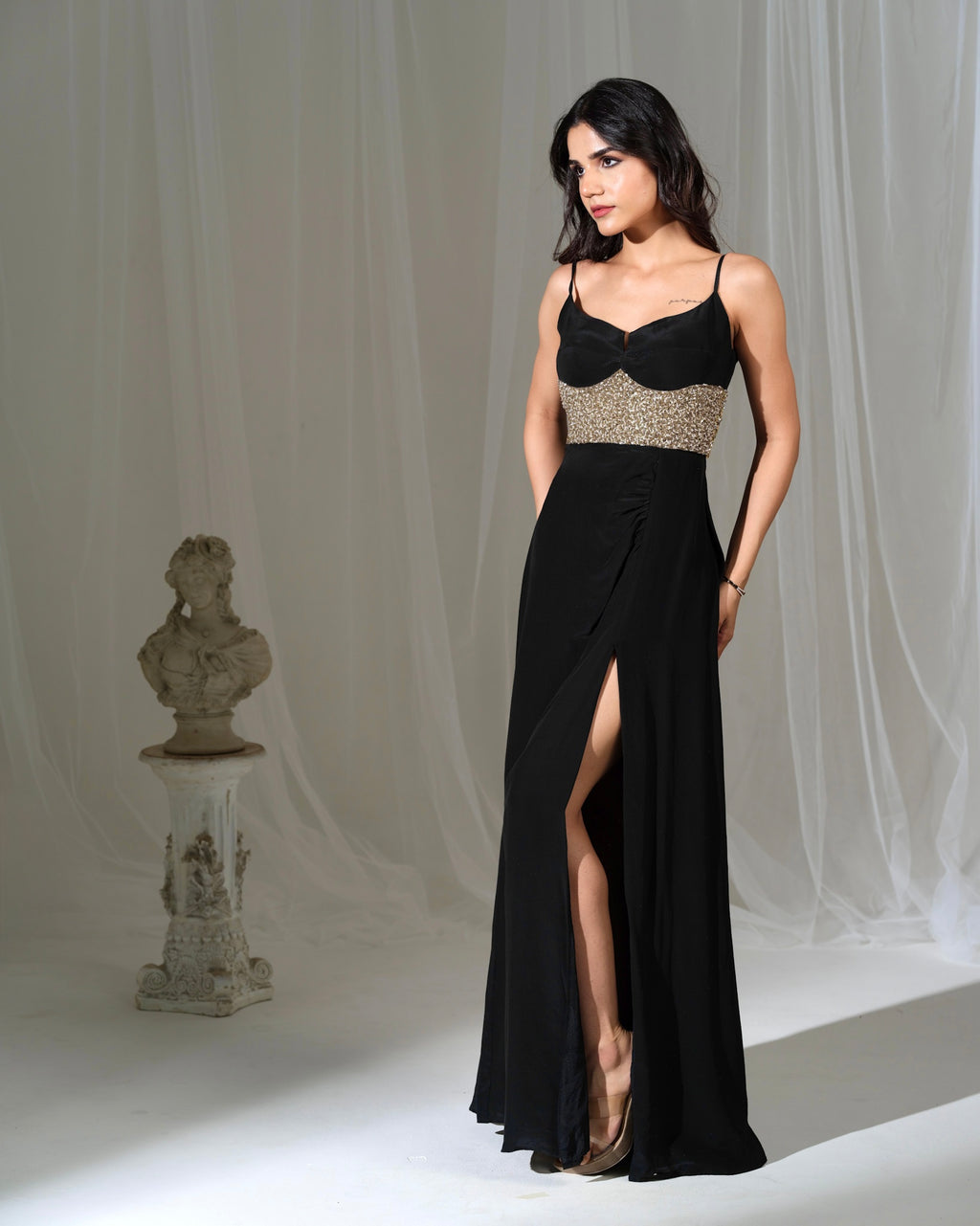Woman in a black evening gown with a high slit standing against a white curtain.