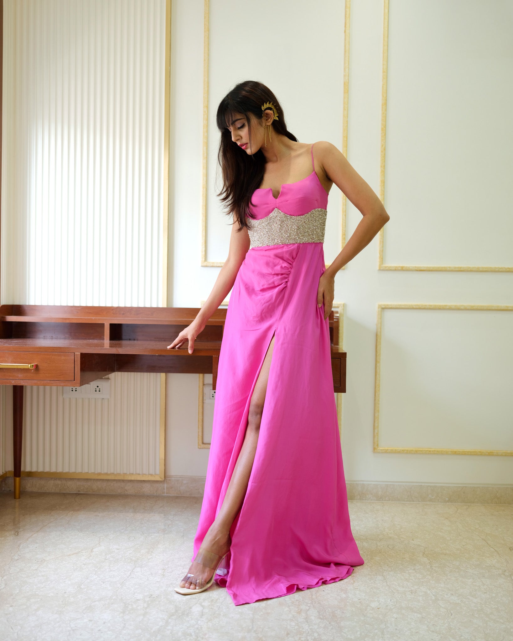 Woman in a pink evening gown with a high slit standing in a room with a desk and wall decorations.