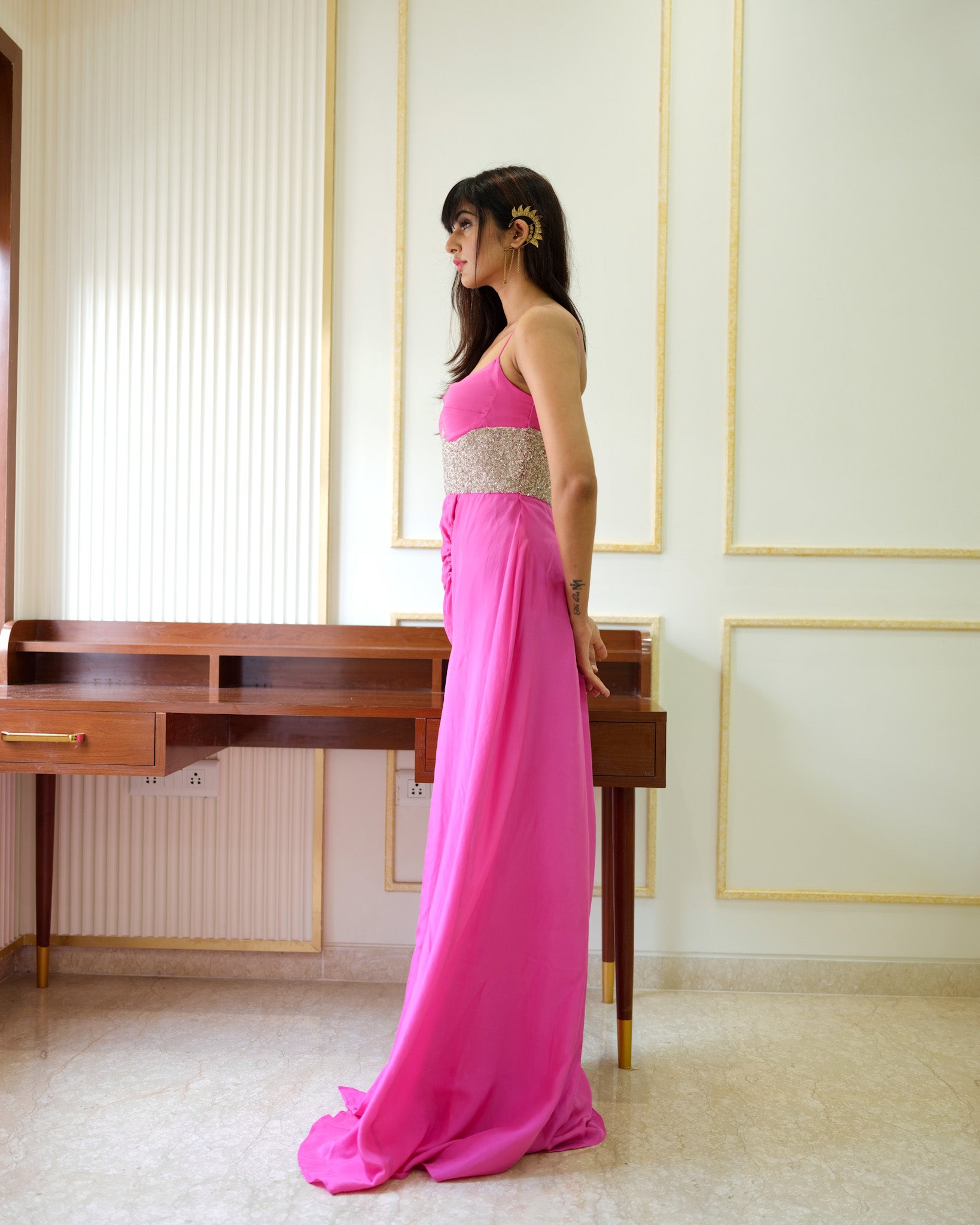 Woman in a pink dress standing in a room with a desk and window blinds.