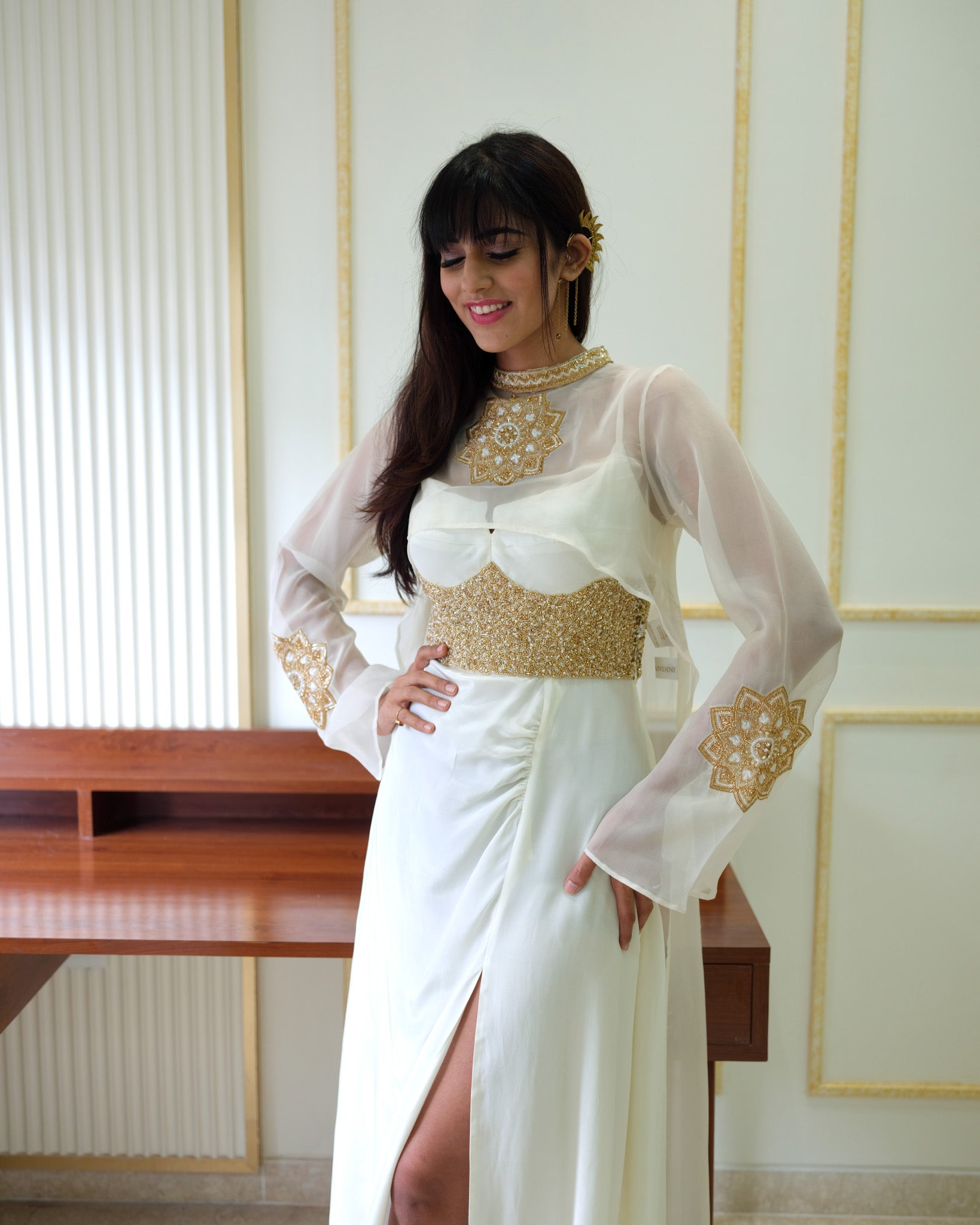 Woman wearing a white and gold dress with a desk and wall in the background