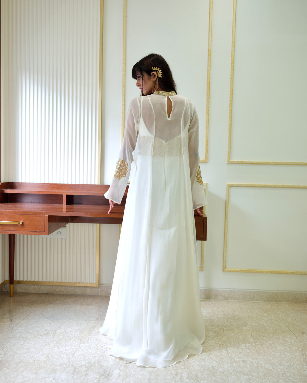 Woman in a white dress standing in a room with a desk and decorative wall.