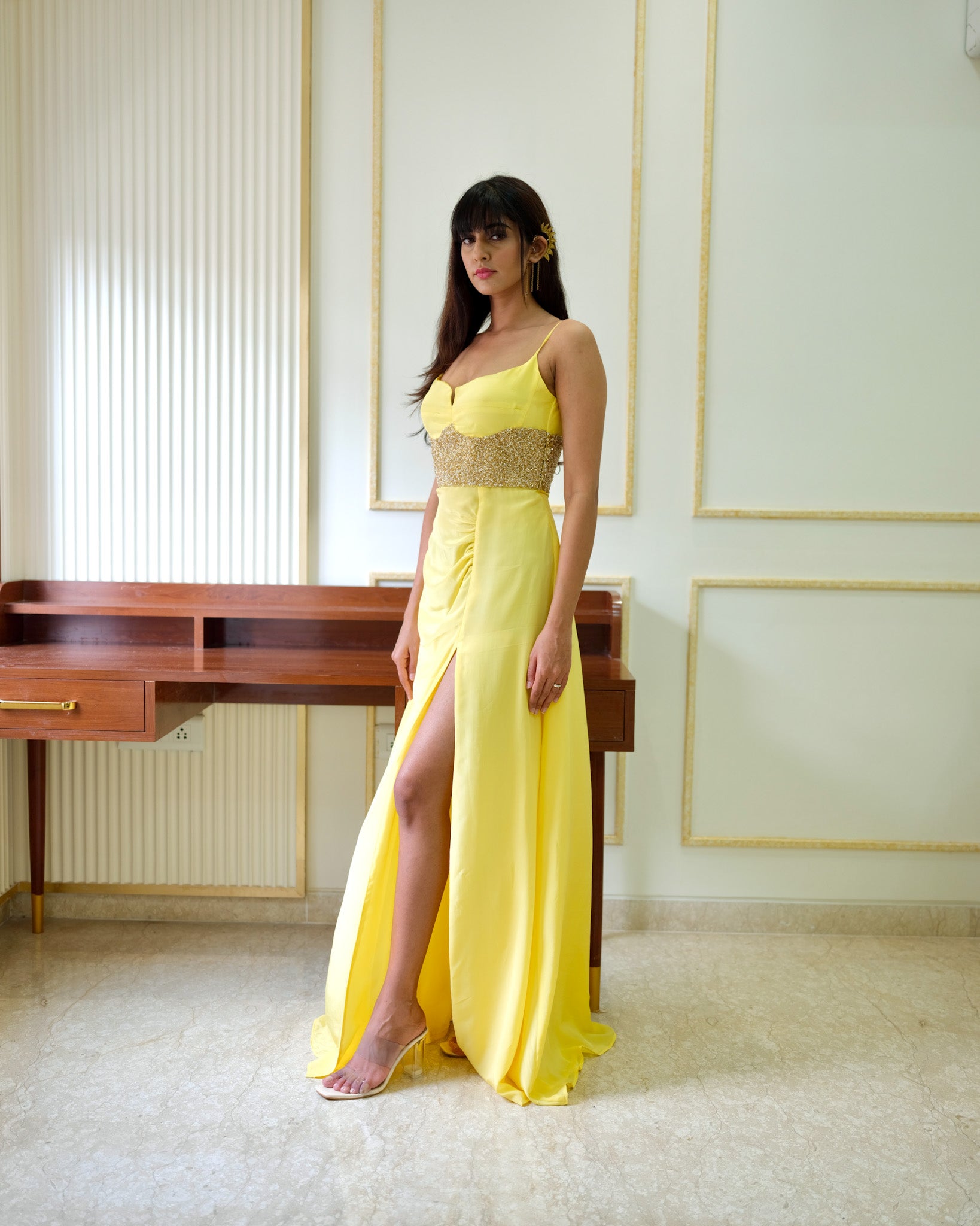 Woman in a yellow evening gown with a high slit standing in an elegant room.