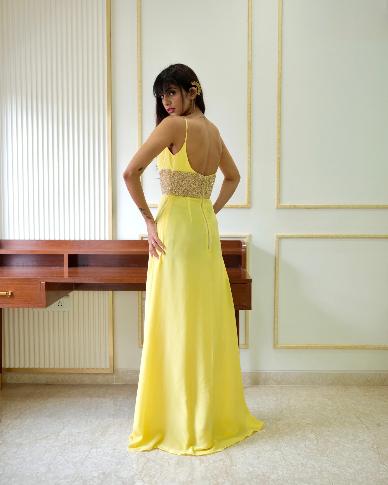 Woman in a yellow evening gown standing in an elegant room with a desk and framed pictures.