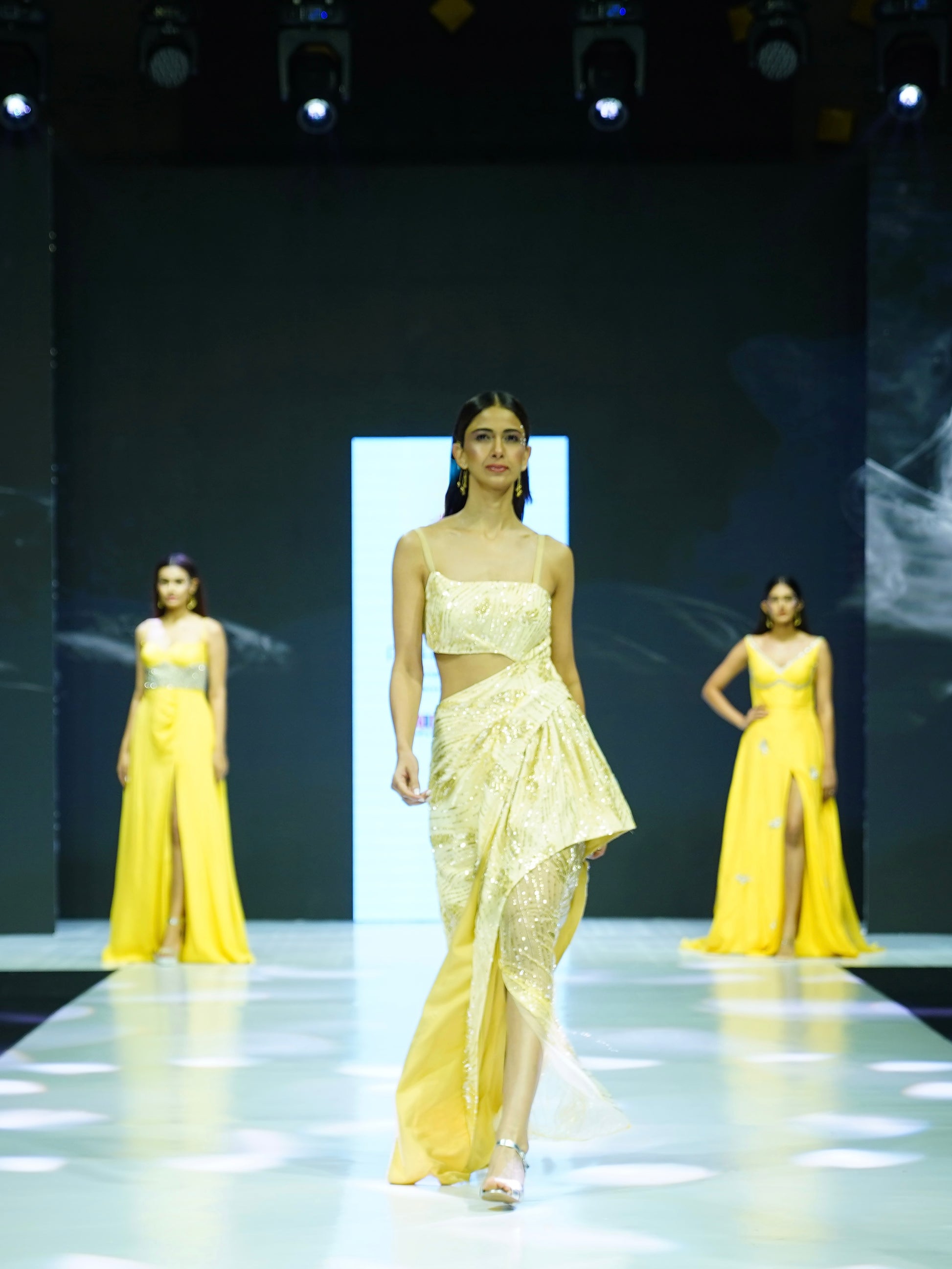 Model walking on a runway wearing a yellow dress with cut-out design.