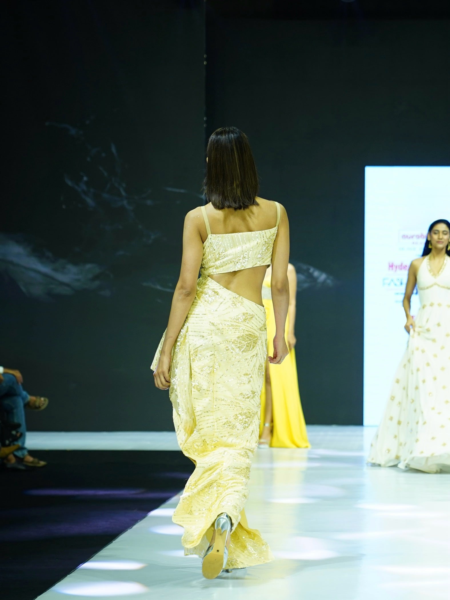 Model walking on a runway wearing a yellow dress with a backless design.