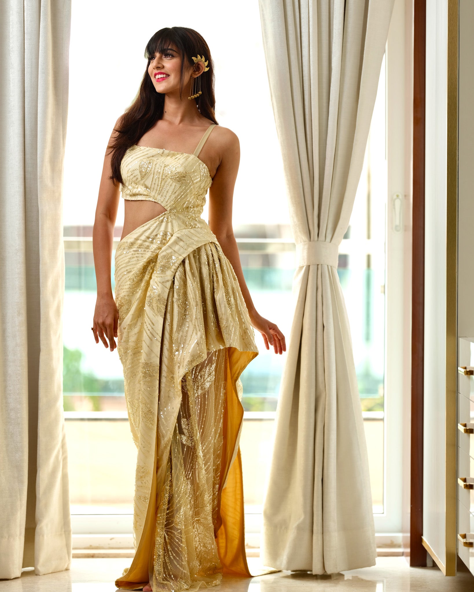 Woman in a gold saree standing in a well-lit room with large windows.