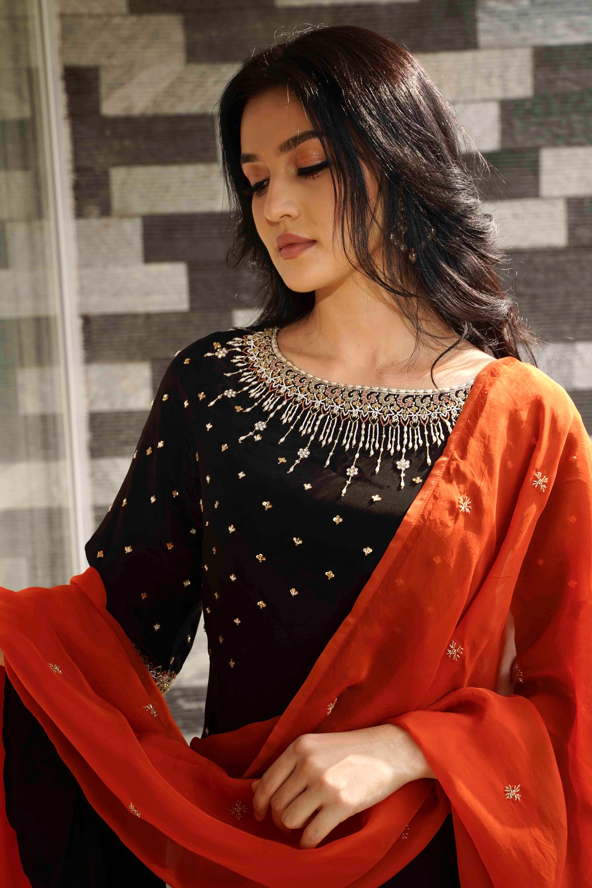 Woman wearing a black top with star patterns and an orange dupatta, against a gray tiled wall.