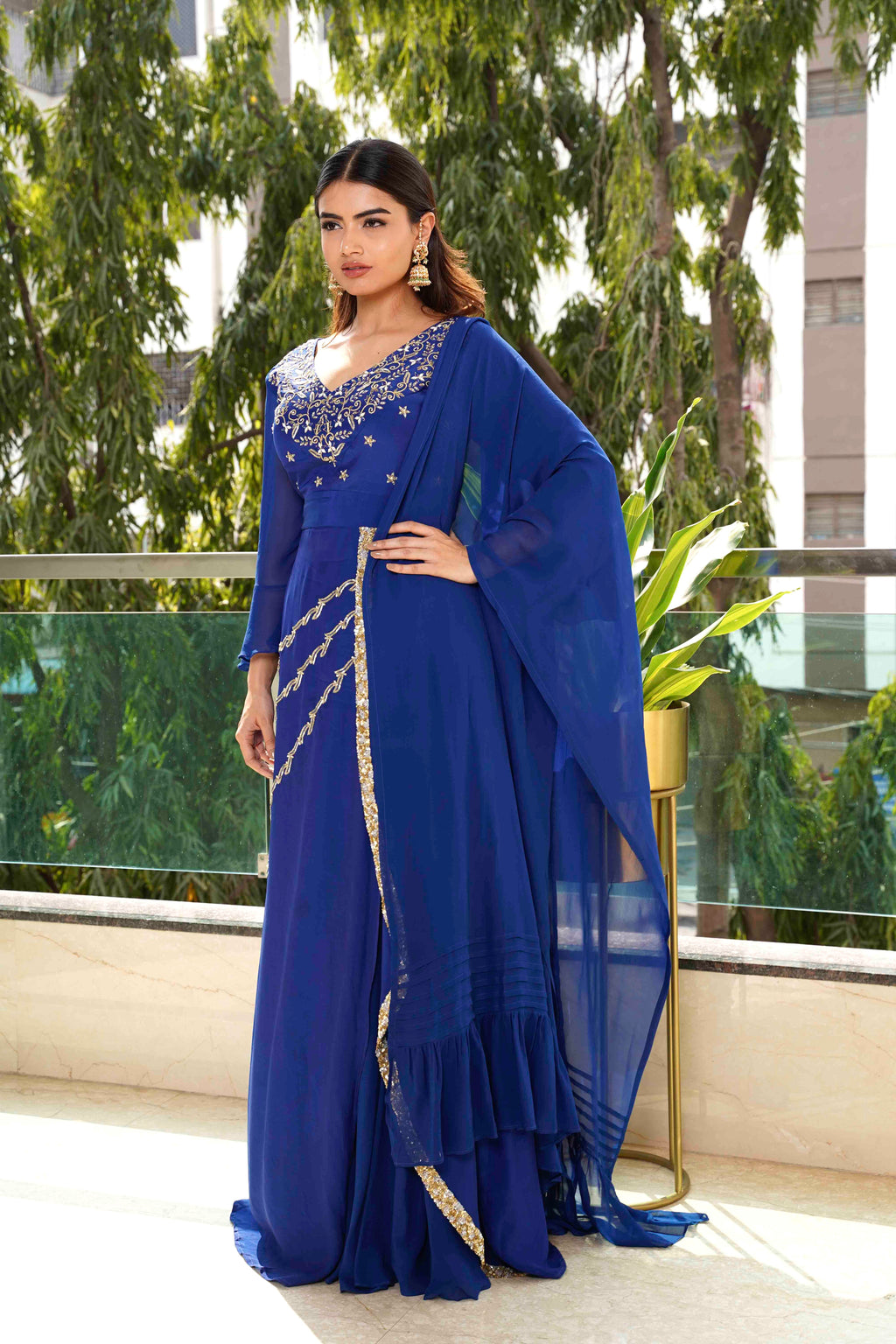 Woman in a blue saree with gold details standing on a balcony with greenery in the background