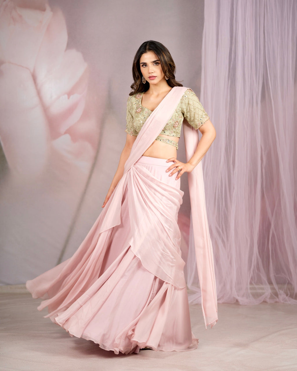 A woman modeling a light pink traditional outfit with a pleated skirt and a matching draped scarf, paired with a light green embroidered blouse, set against a soft pink studio backdrop with sheer drapes.