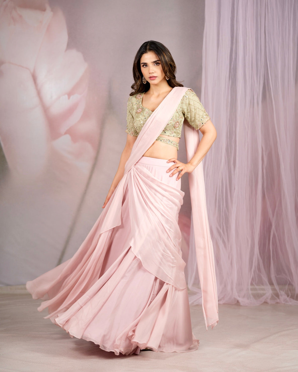 A woman modeling a light pink traditional outfit with a pleated skirt and a matching draped scarf, paired with a light green embroidered blouse, set against a soft pink studio backdrop with sheer drapes.