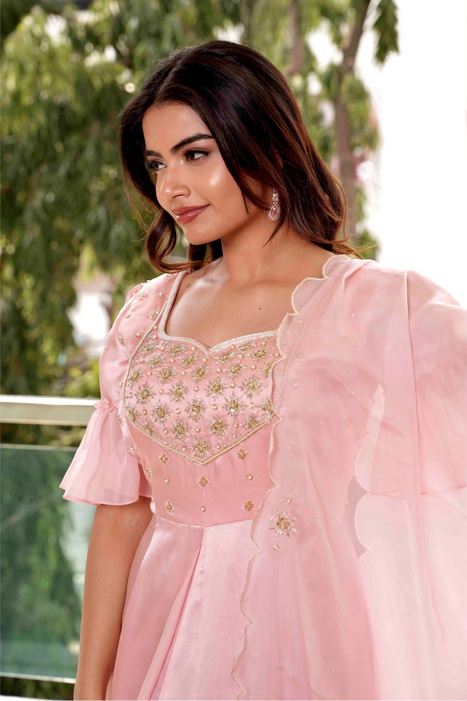 Woman wearing a pink traditional outfit with intricate designs, standing outdoors with greenery in the background.