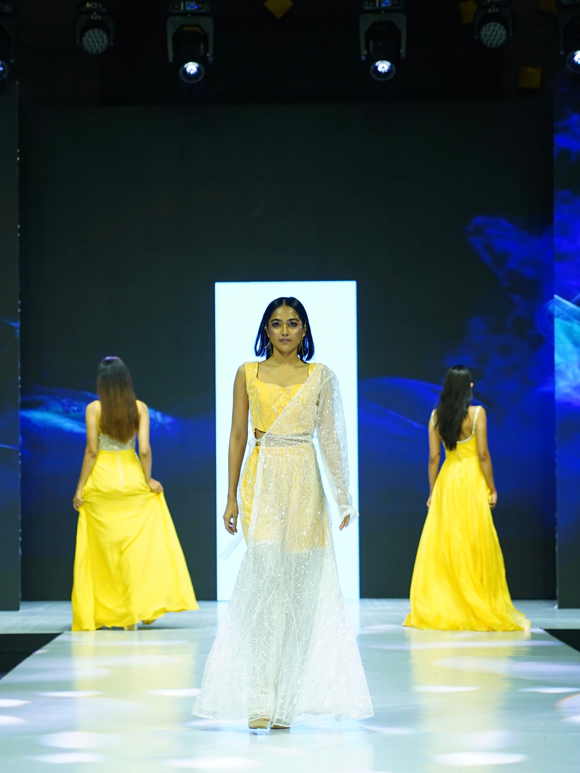 Model walking down a runway wearing a yellow and white dress with two other models in yellow dresses on either side.