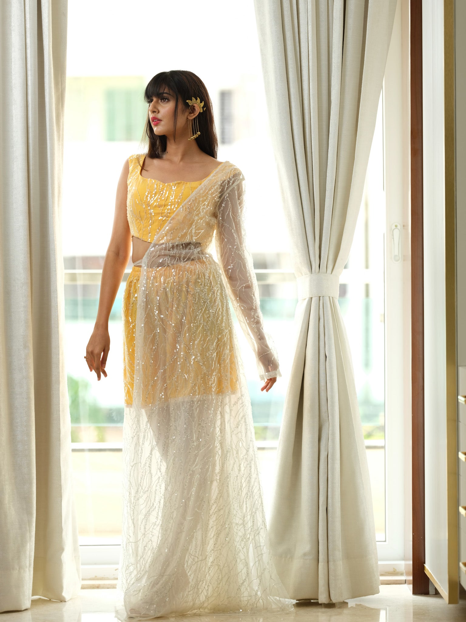 Woman in a yellow and white traditional outfit standing in a bright room with large windows.