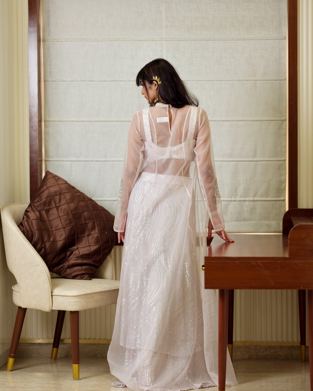 Woman in a white dress standing in a room with a chair and table.