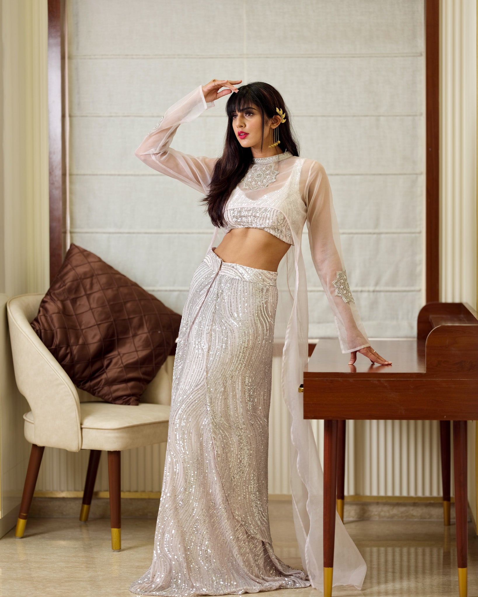 Woman in a sparkling silver outfit posing in a room with a chair and table.