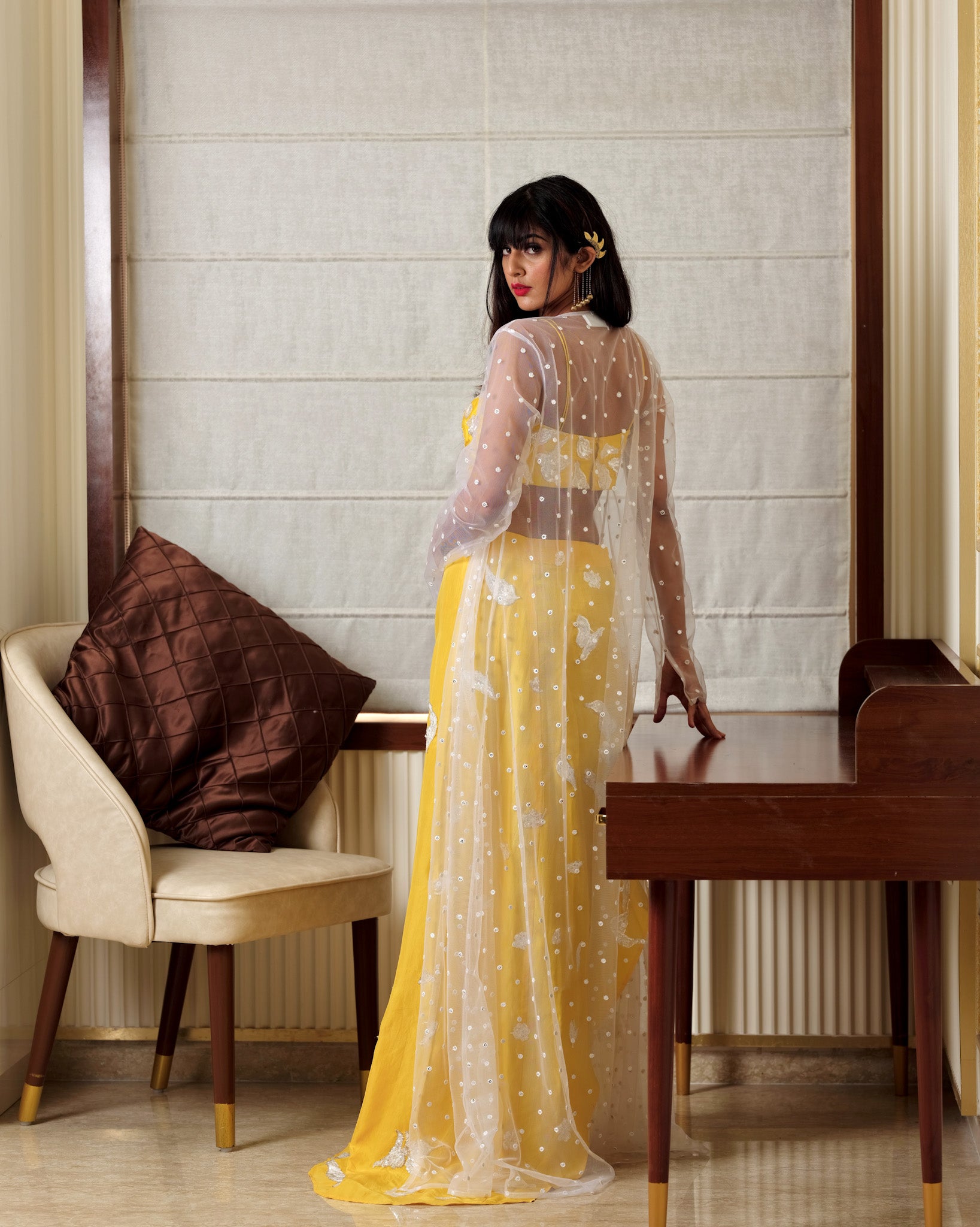 Woman in a yellow and white dress standing in a room with a chair and table.
