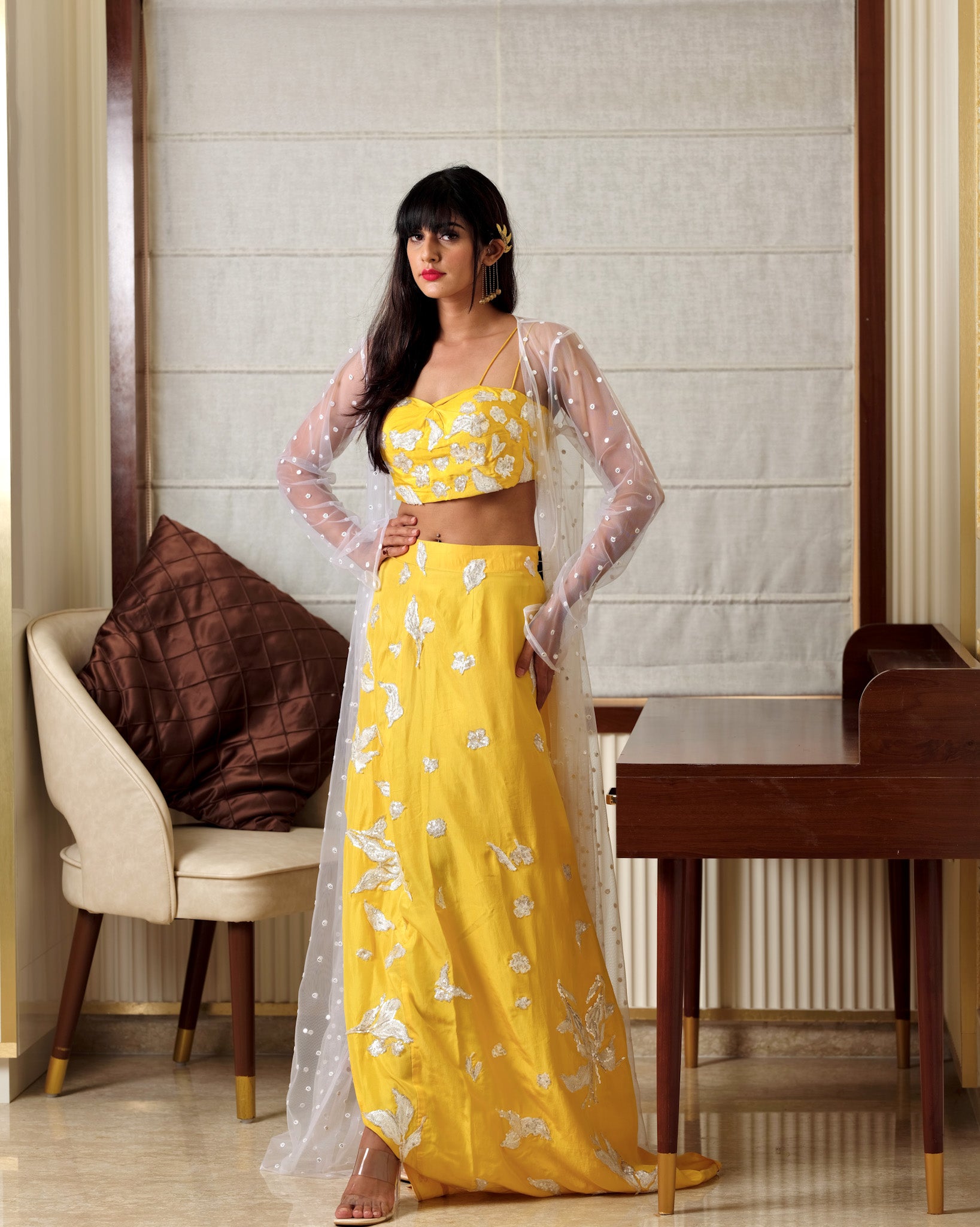Woman in a yellow floral outfit standing indoors with a chair and table in the background.