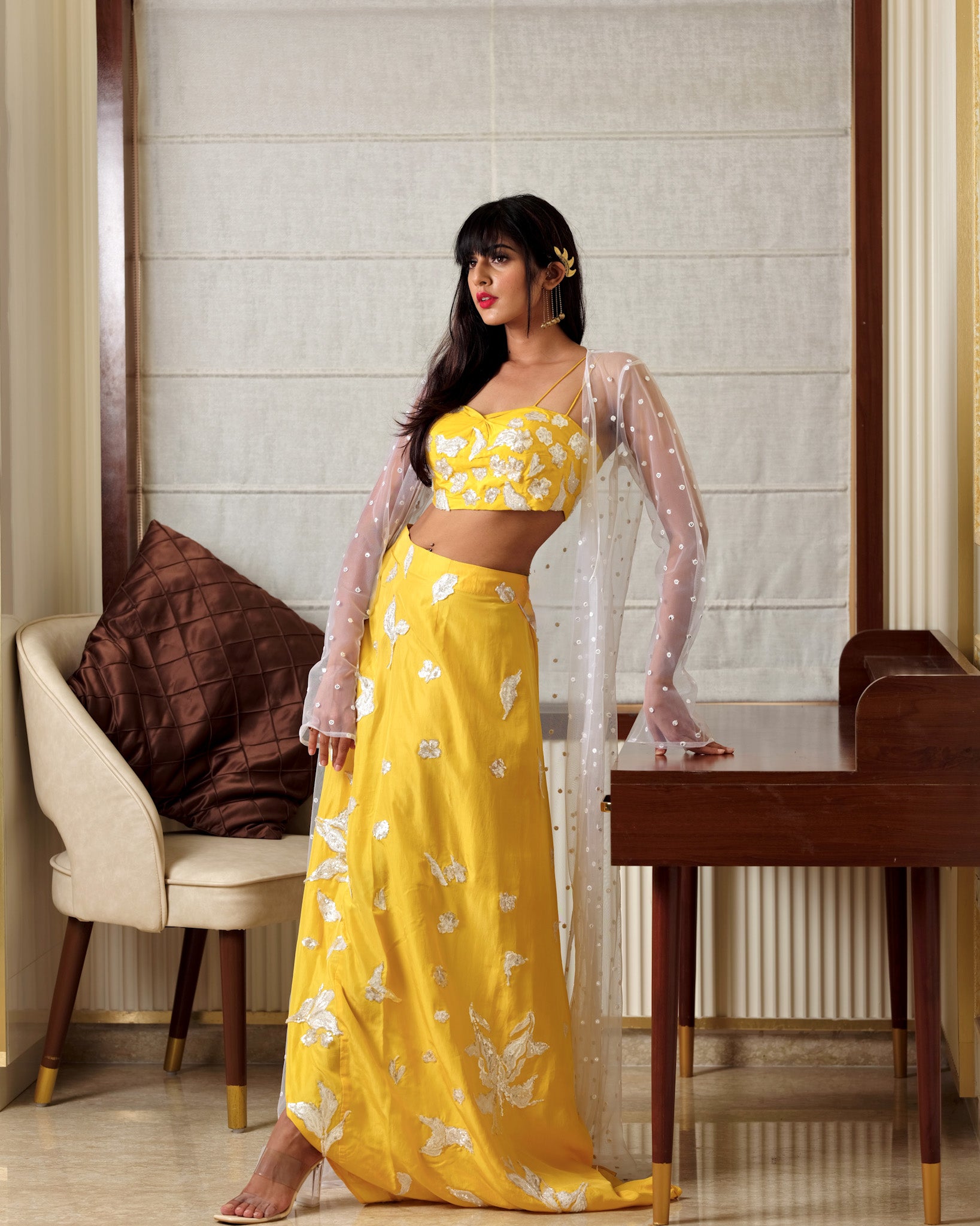 Woman in a yellow floral outfit standing in a room with a chair and table.