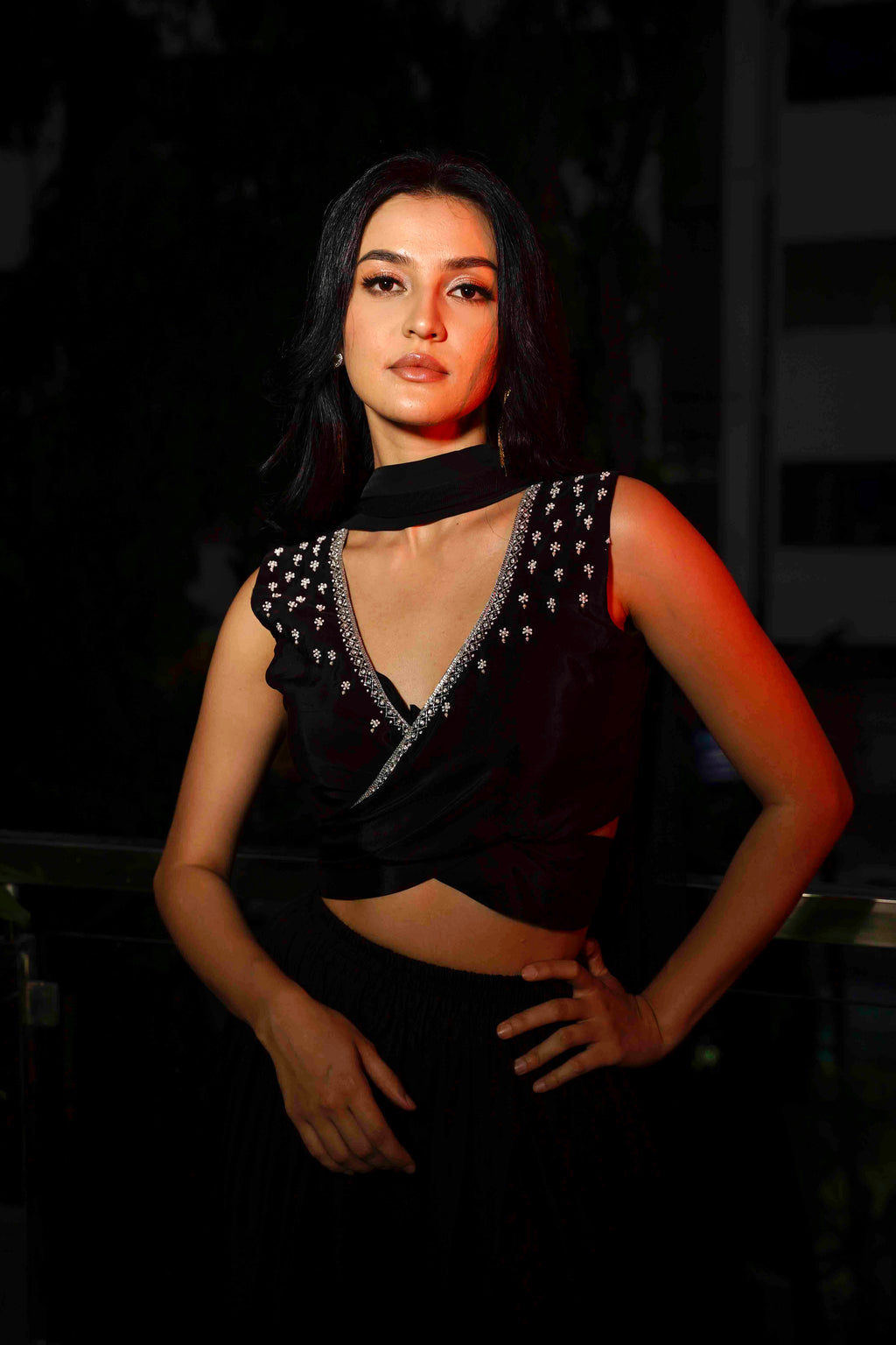 A woman modeling a black outfit with a structured cross-belt wrap blouse and voluminous pants, accessorized with a soft tulle dupatta.