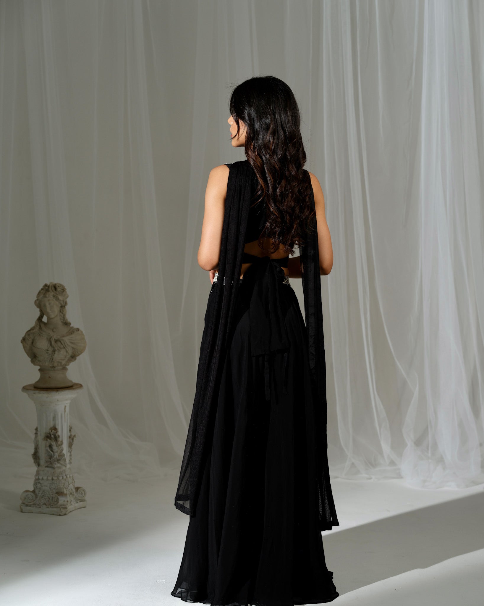 Woman in a black dress standing in a studio with white curtains and a bust statue.