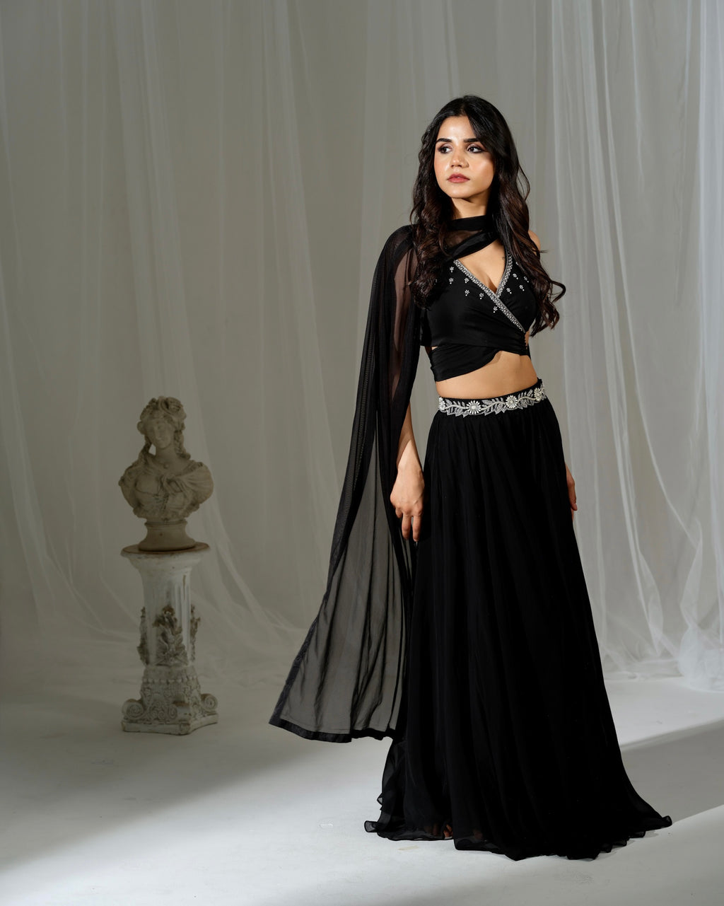 Woman in a black outfit with sheer overlay standing against a white curtain.