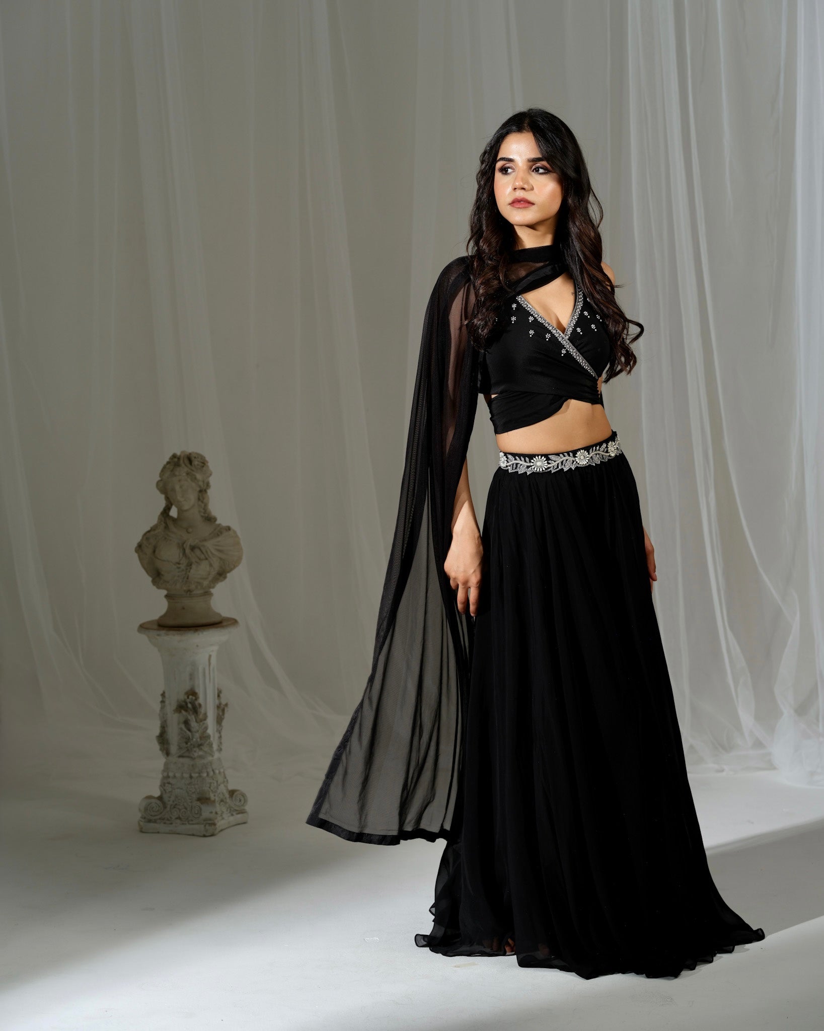 Woman in a black outfit with sheer overlay standing against a white curtain.