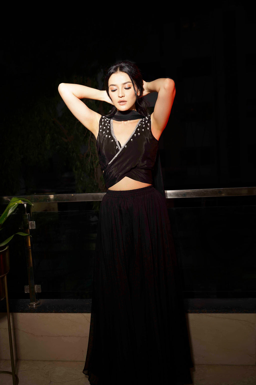 A woman modeling a black outfit with a structured cross-belt wrap blouse and voluminous pants, accessorized with a soft tulle dupatta.
