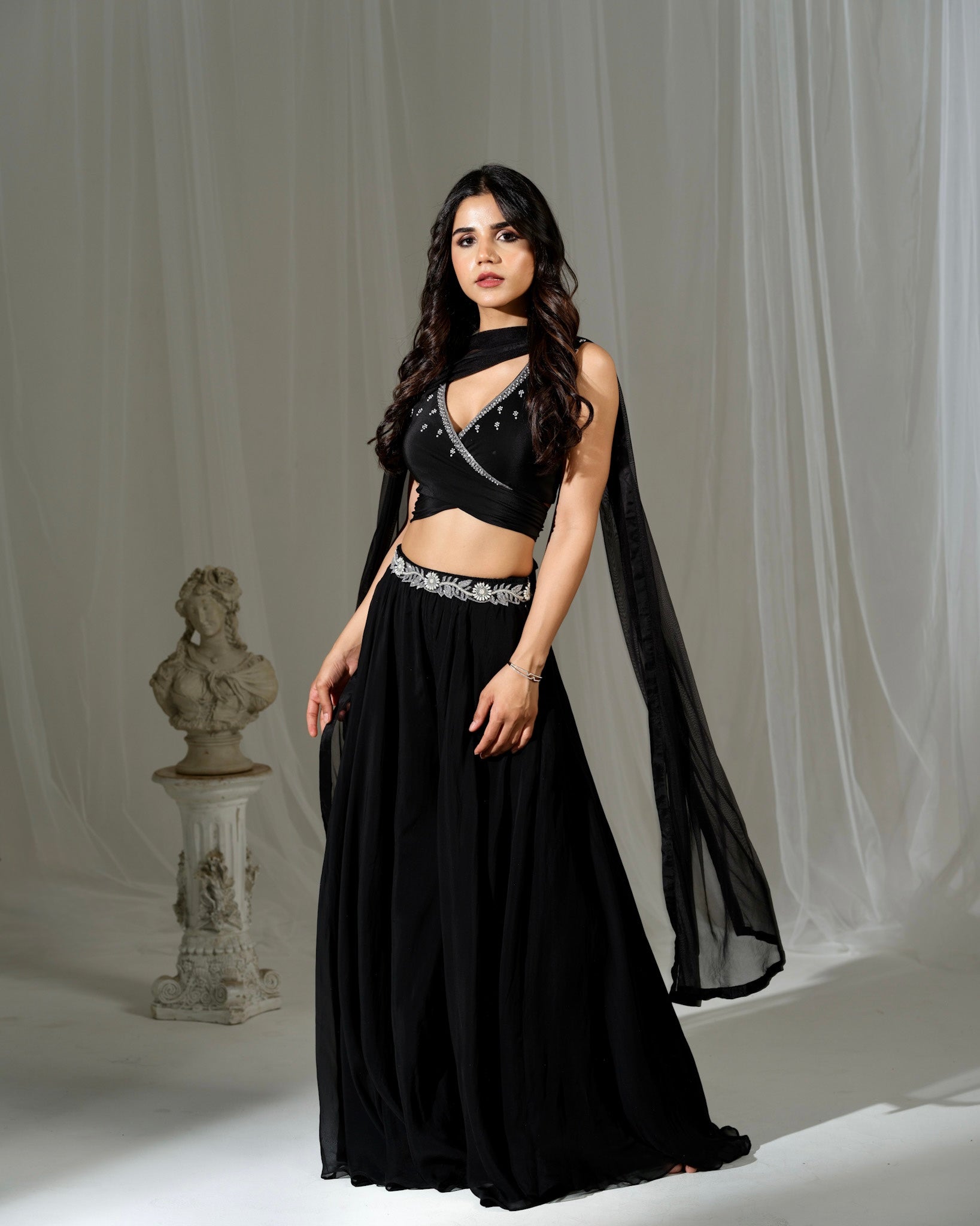 Woman in a black outfit with a sheer cape standing against a white curtain.