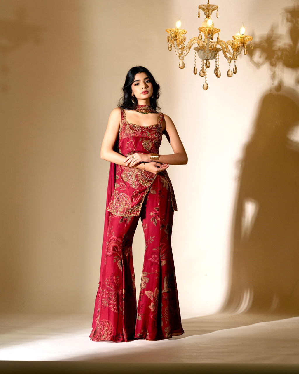 Woman in a red saree with gold patterns standing in front of a chandelier.
