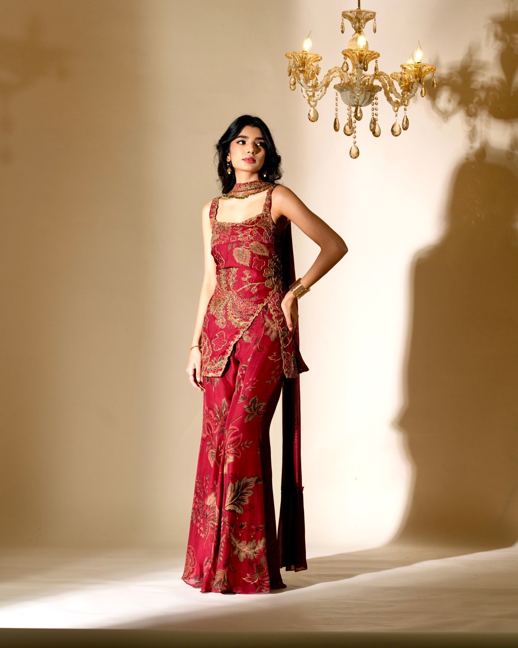Woman in a red patterned dress standing in a softly lit room with a chandelier.