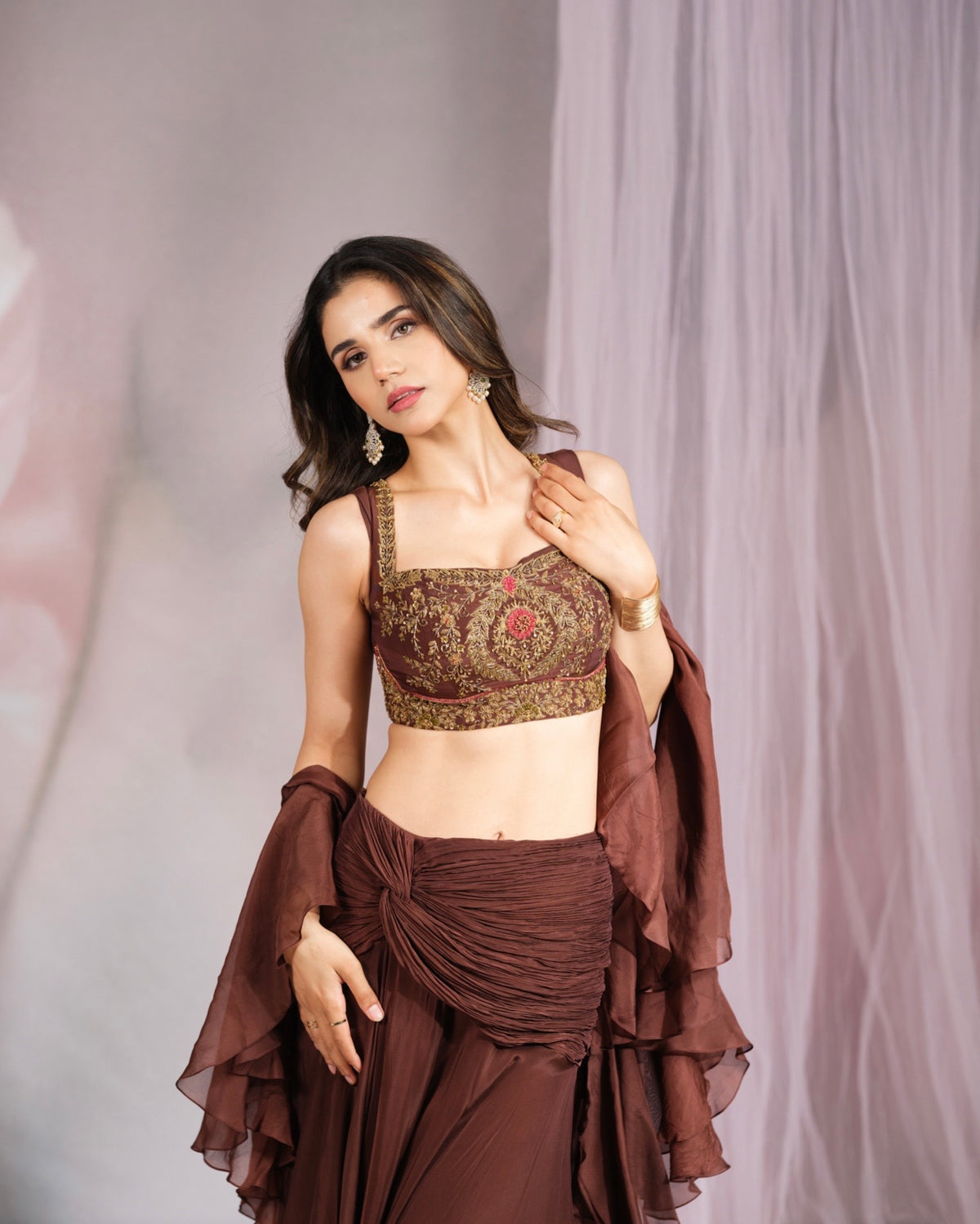 A woman models a brown embroidered crop top and matching draped skirt set against a soft, draped lavender backdrop.
