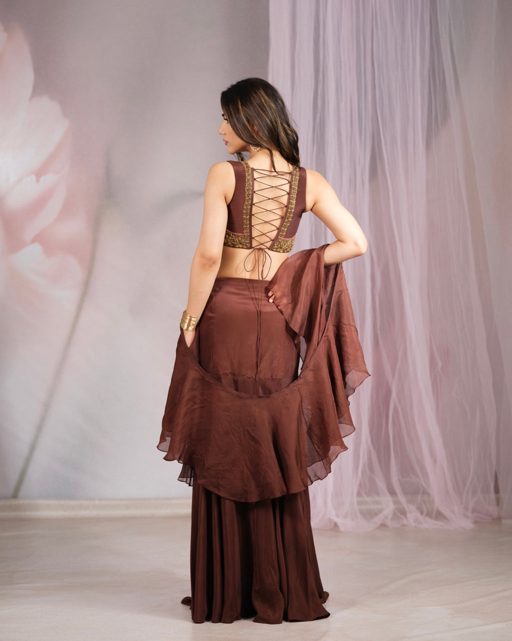 A woman shown from the back wearing a brown traditional outfit with a lace-up open back and a draped sheer matching fabric, standing in a softly lit studio with pale pink tulle curtains.