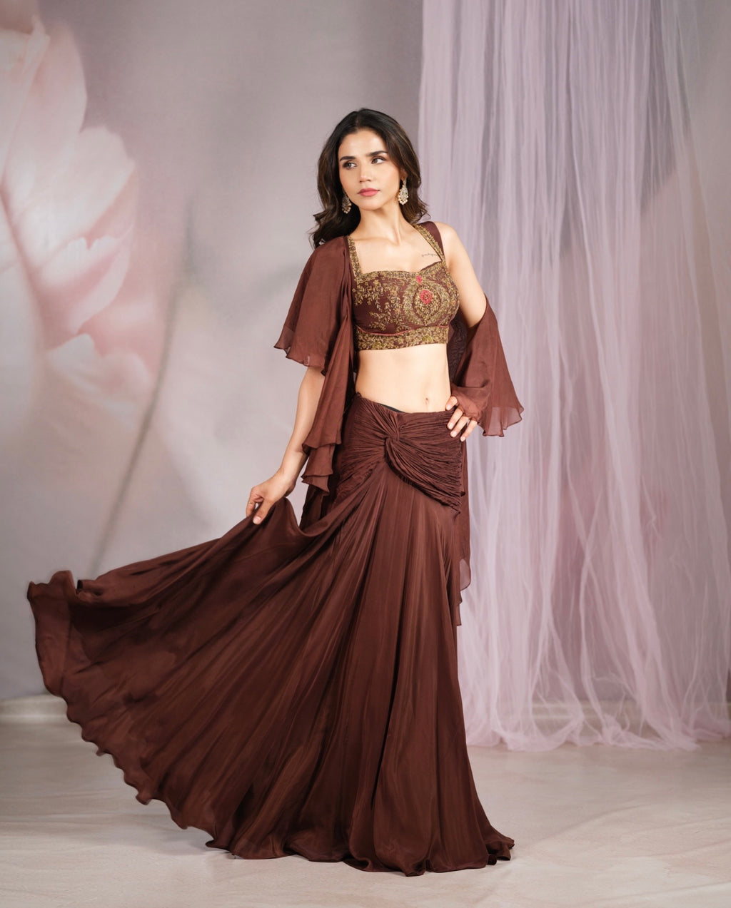 A woman models a brown traditional Indian outfit consisting of a pleated maxi skirt, a matching draped blouse, and an embroidered crop top. She is standing in a studio with a soft pink floral backdrop and sheer white drape.