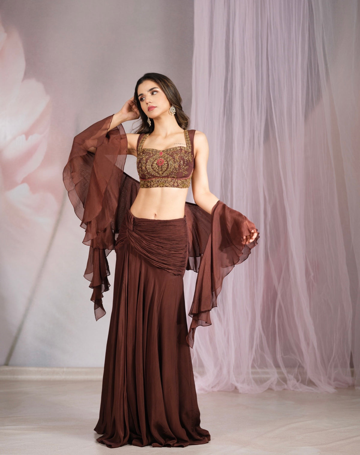 A woman models a brown two-piece traditional outfit with a heavily embroidered crop top, a draped maxi skirt, and sheer flutter sleeves against a soft pink and white backdrop.