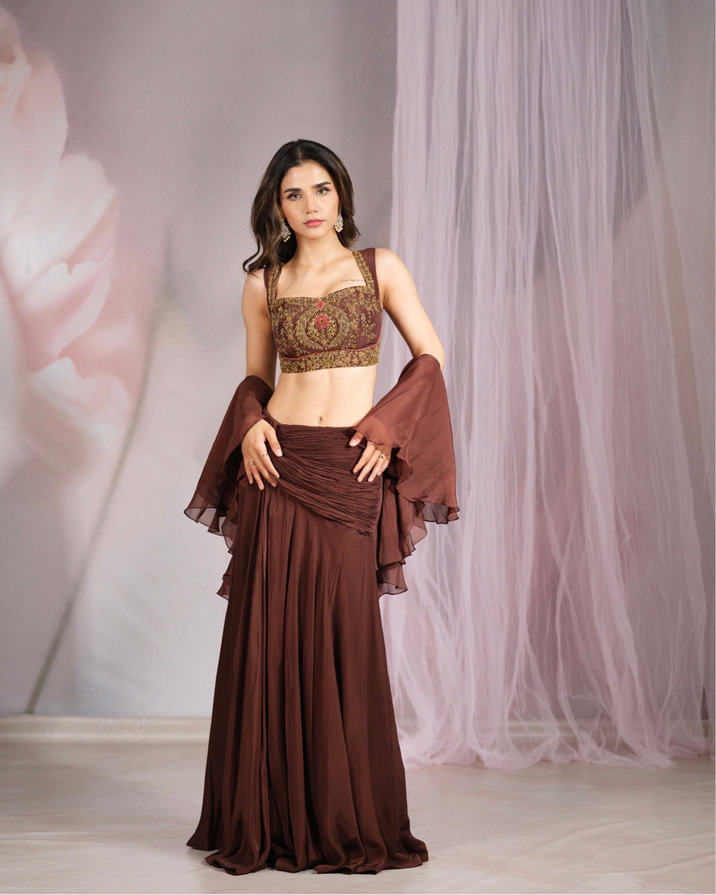A woman models a brown traditional Indian outfit consisting of a heavily embroidered crop top, a matching draped skirt, and sheer flutter sleeves, set against a soft pink and white tulle backdrop.