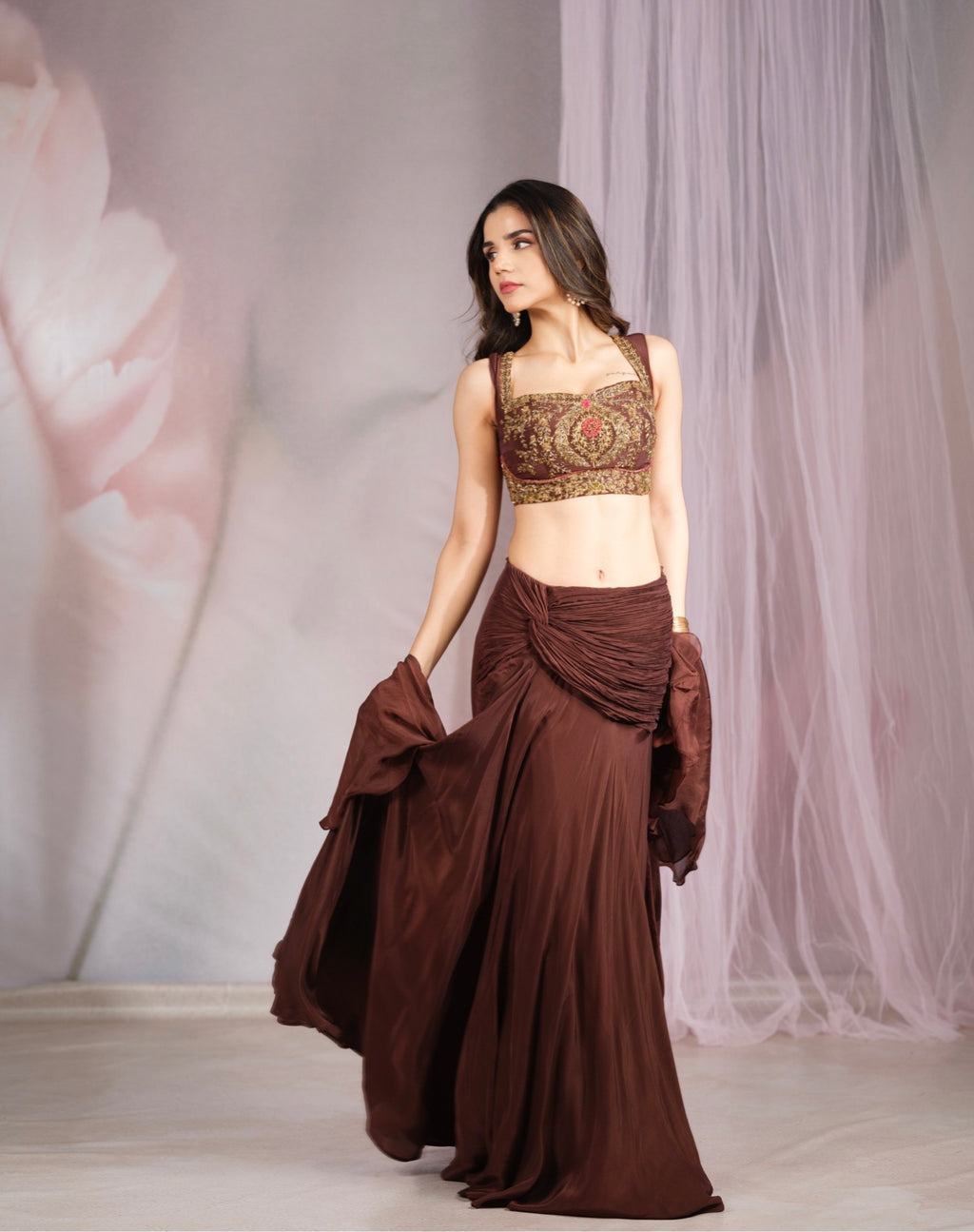 A woman modeling a brown traditional outfit featuring an embroidered crop top and a draped maxi skirt, standing against a soft pink and white backdrop.