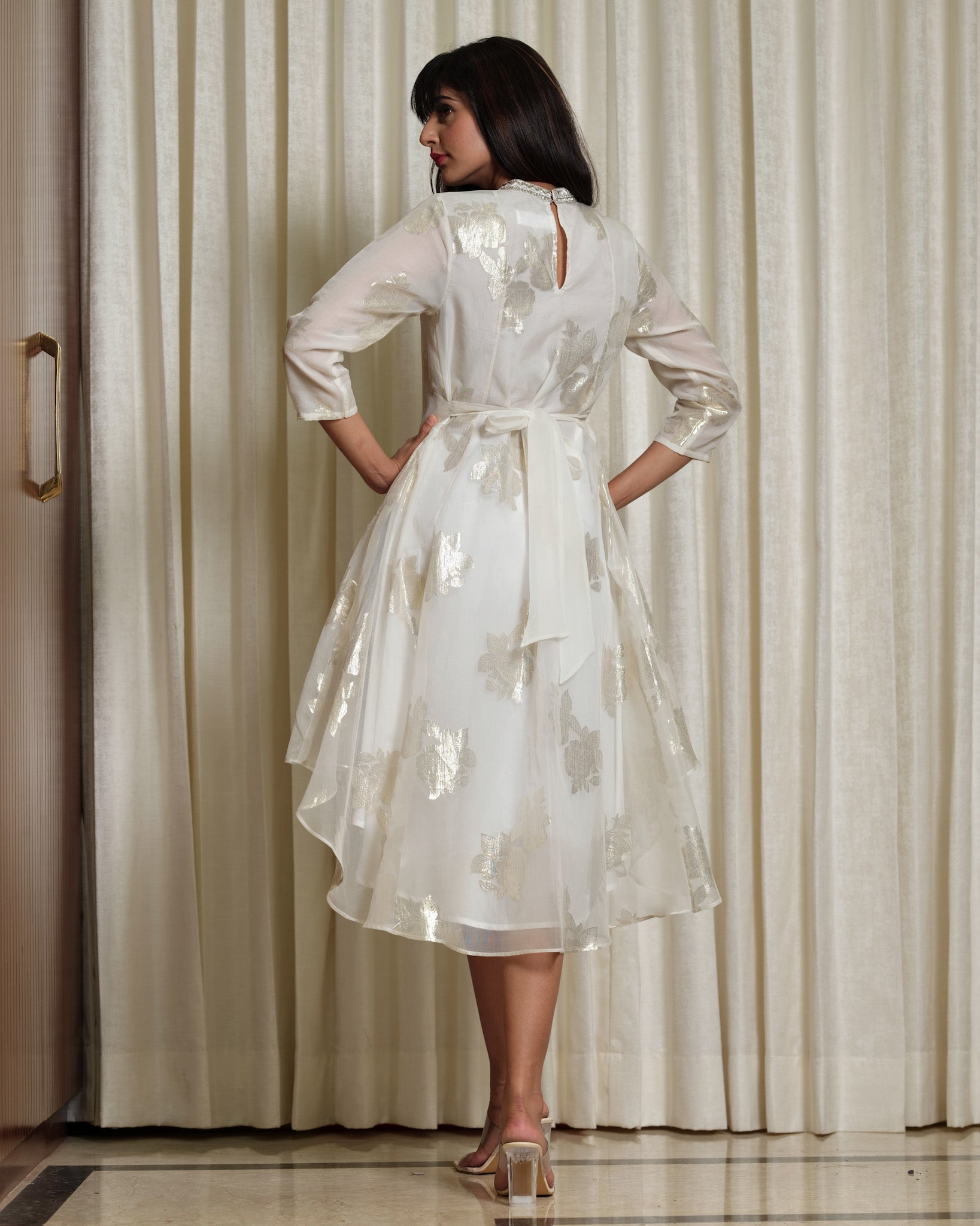 Woman wearing a white dress with floral patterns in front of a curtain.