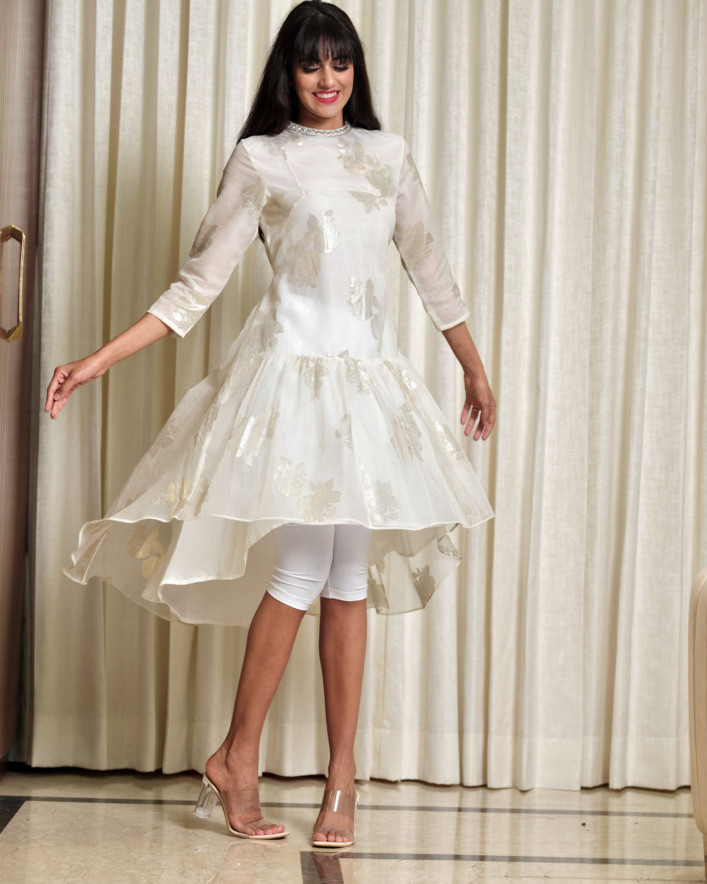 Woman wearing a white dress with a high-low hem in front of a curtain.