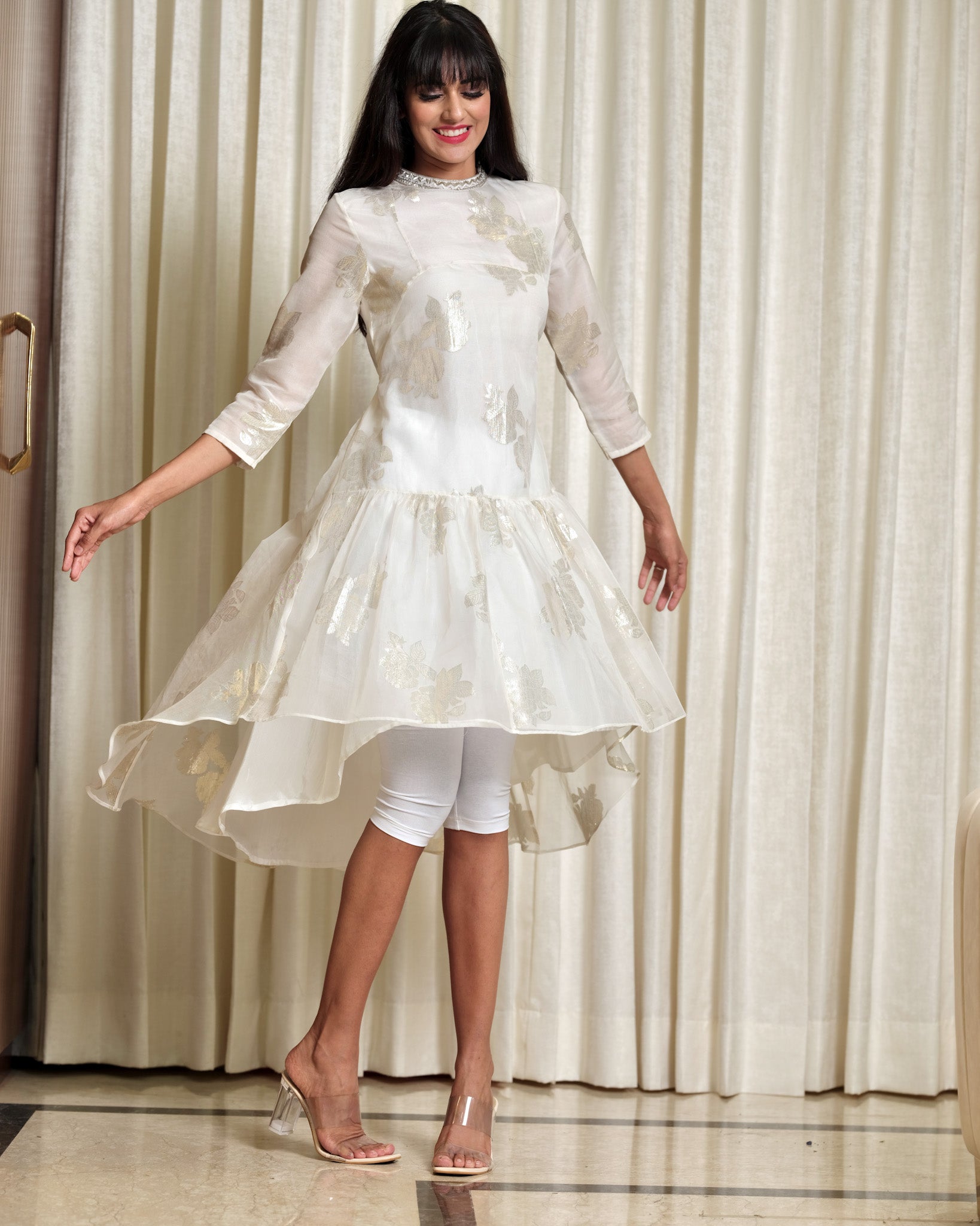 Woman wearing a white dress with a high-low hem in front of a curtain.