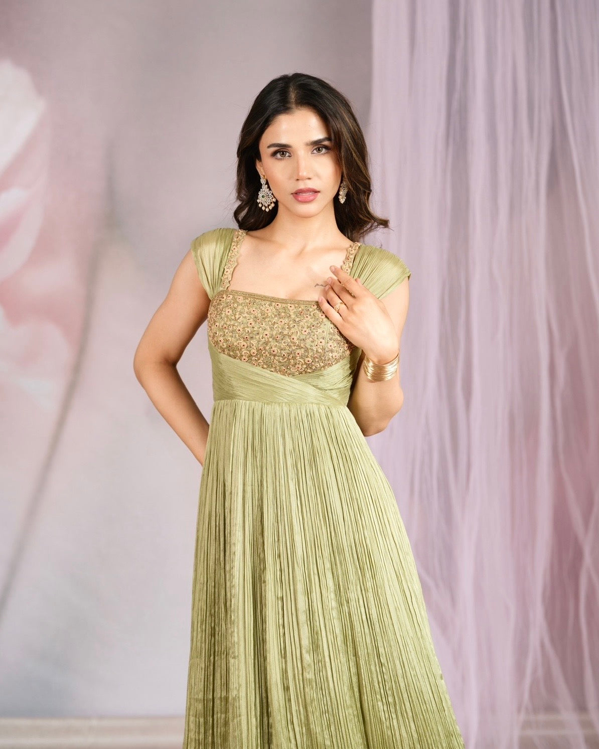 A woman modeling a light olive green sleeveless gown with a heavily embellished gold sequined bodice and a pleated skirt, set against a soft pink and white draped studio backdrop.