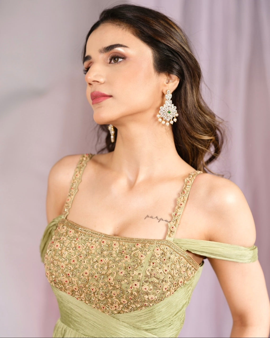 A woman wearing a light olive green off-the-shoulder dress with intricate floral embroidery on the bodice, paired with statement earrings, set against a soft lavender background.