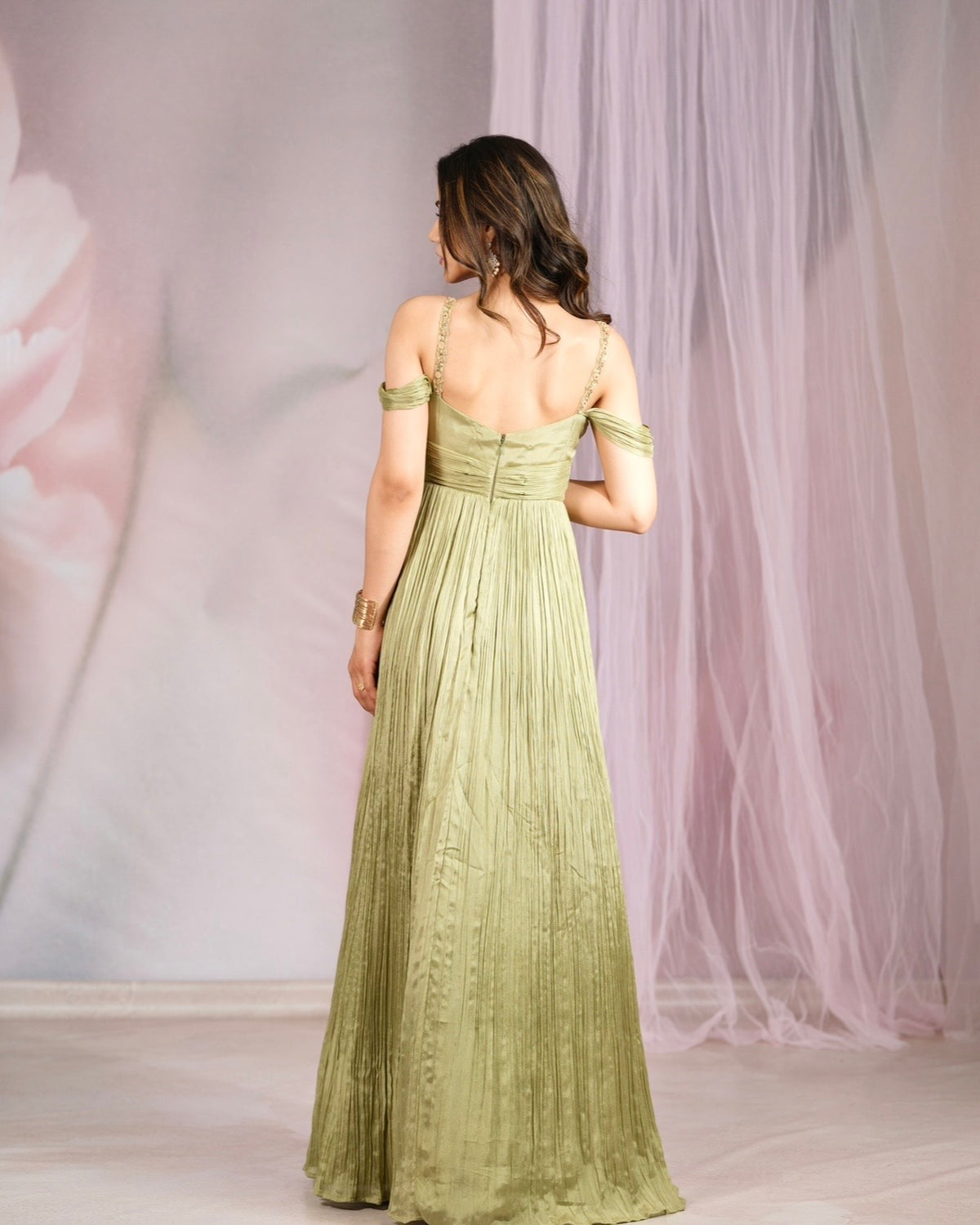 A woman viewed from the back wearing a floor-length off-the-shoulder olive green dress with a pleated skirt, standing in front of a soft pink and white draped backdrop.
