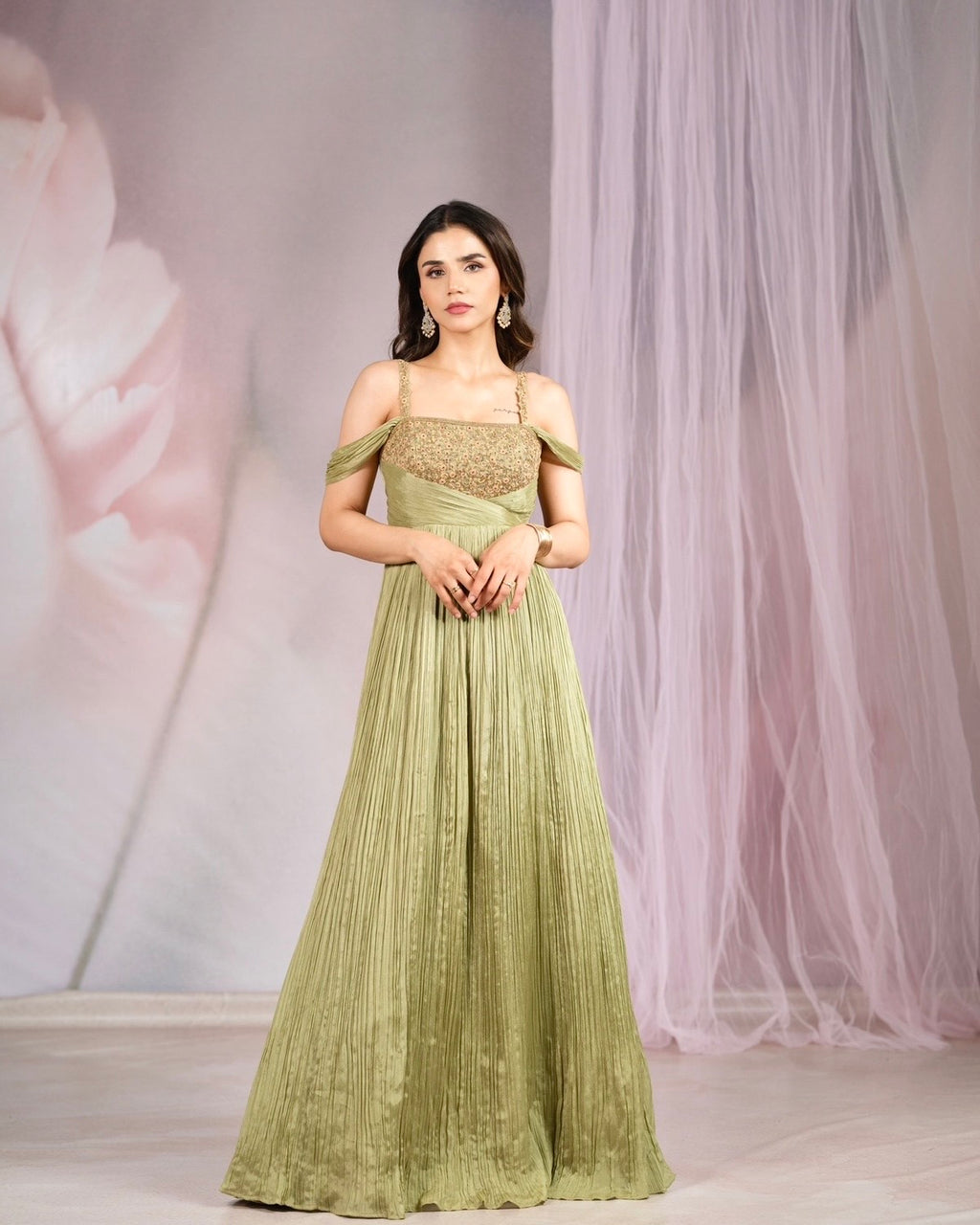 A woman models a floor-length olive green evening gown with a textured, embroidered bodice and off-the-shoulder straps, set against a soft pink floral backdrop with sheer tulle drapes.